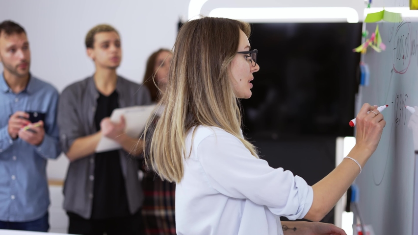Young woman ofice worker standing and writing on white board an talking with her colleagues