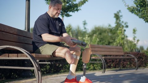 Mature Caucasian amputee putting on prosthesis in slow motion sitting on bench talking. Wide shot grey-haired handsome confident man in sunny summer park outdoors with artificial limb.