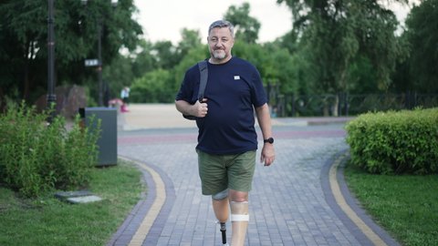 Front view portrait confident grey-haired male amputee walking in slow motion in summer park looking around. Wide shot positive Caucasian man with grey hair and prosthetic legs strolling outdoors.