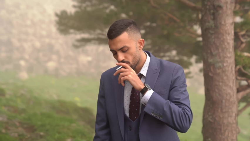 a man with a beard smokes a cigarette standing in the woods by a tree. smoke break at work. bad habit and dependence on nicotine. businessman.