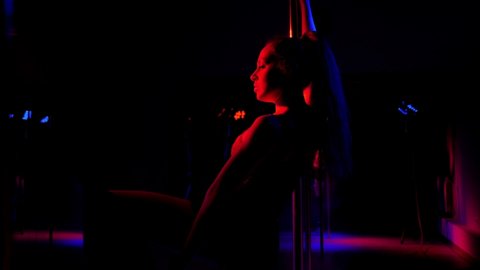 The silhouette of a woman dances a sensual dance on the floor near a pole in the dark under the light of bright spotlights.