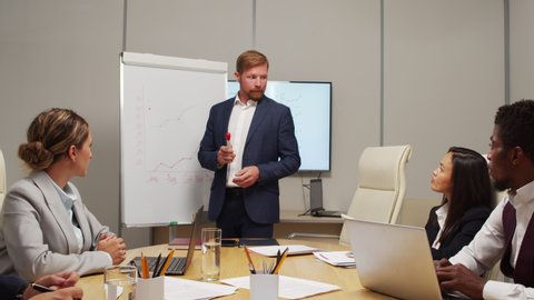 Dolly-out shot of businessman in suit standing before whiteboard and delivering financial report to team of colleagues sitting around table in meeting room