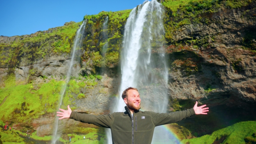 Man Posing Happily For The Camera With A Rainbow On The Seljalandsfoss Waterfall In South Region Of Iceland. wide shot