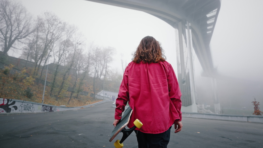 Back view of a young teenager with a skateboard under his arm. A long-haired trendy man wearing a red shirt walks through the city park in the background near the bridge. Foggy morning.
