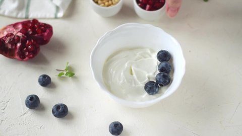 Adding pomegranate seeds into Greek yogurt with blueberry in the white ceramic bowl. Thick dairy product with topping. Making healthy breakfast