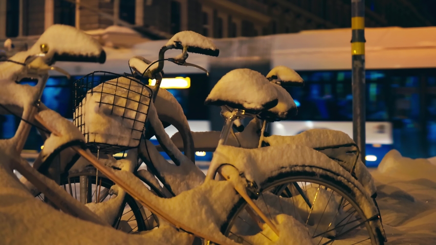 Two bicycles covered in snow parked in city center with traffic and people in background
