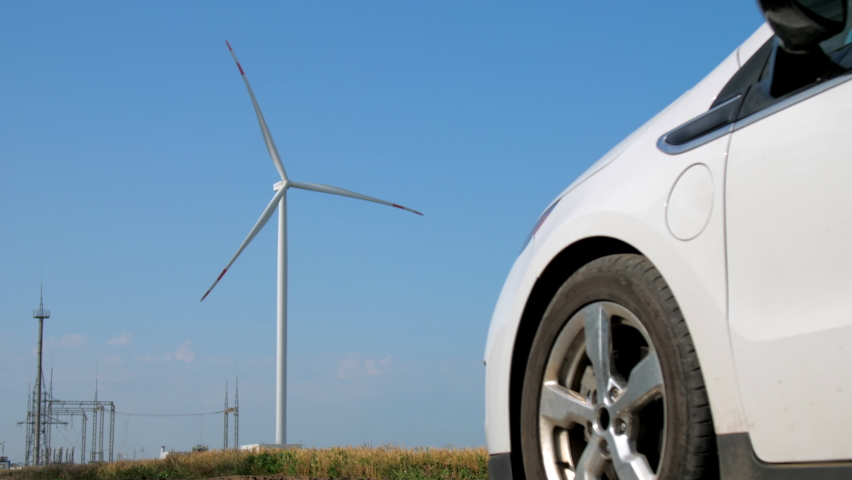 Windmill generates alternative energy in field. Man charges white electric car against rotating propeller near electrical transmission lines Royalty-Free Stock Footage #1083340420