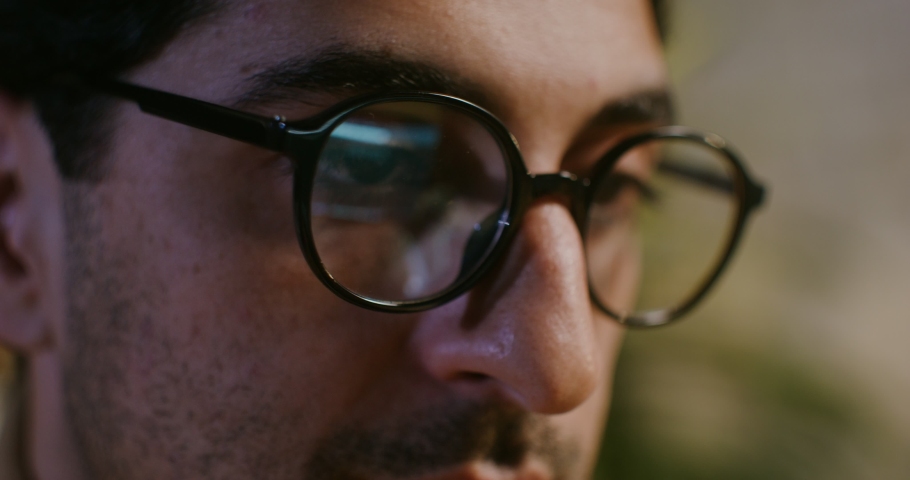 A young brunette man in glasses with stubble on his face looks intently at the laptop screen while typing a text, a close-up of the face