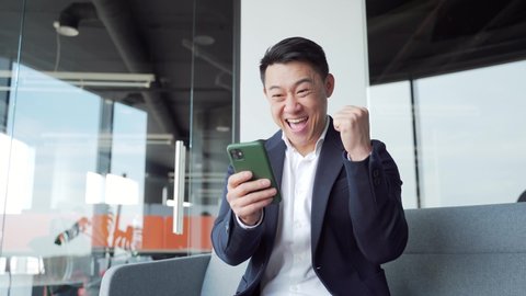 happy euphoric asian business man wear suit celebrate mobile win, excited overjoyed lucky executive winner receive good news in message use smartphone rejoice bet financial success victory in office