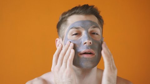 Caucasian man with a gray clay mask on his face poses in front of the camera on an isolated yellow background - the concept of beauty and skin care.