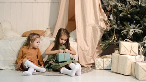 Children sitting on the floor by the bed and decorated Christmas tree with Christmas gifts unwrap one of the gift boxes. Children received gifts for xmas from Santa. Celebrating the New Year