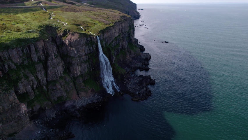 Aerial shot of flowing waterfall on the side of a cliff into ocean, shot in Iceland. Green grass and birds flying below.