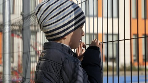 Upset and stressed boy looking through school metal fence after was bullied. Concept of poverty, immigration, bullying and kids stress