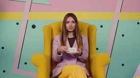 Stylish bossy woman is sitting in yellow armchair in creative space, drinking coffee from white cup and looking at camera, confident and successful lady, Slow motion.