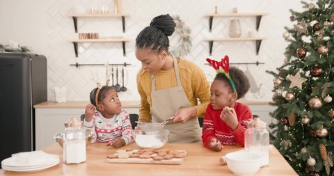 Joyful African American mom with little cute kids standing at table in Christmassy decorated kitchen and having fun while making xmas cookies at home. New Year preparations. Holidays concept