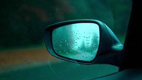 Close-up, side rearview mirror of driving car in autumn rainy day. water drops on rear view mirror of moving automobile. Selective focus. Travel by car. Green trees, clouds over gray road