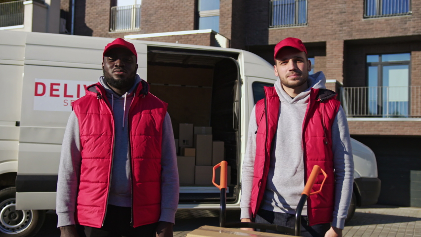 African american and caucasian couriers standing in front of delivery van with parcel boxes on a trolley. Delivery men cross their hands, looking at the camera.
