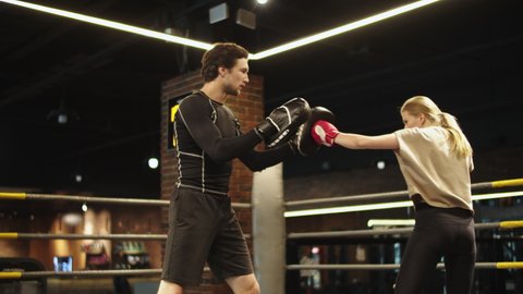 Serious male boxer instructing female boxer on boxing ring. Young fit girl doing punches in sport club. Strict coach holding punching pads at gym.