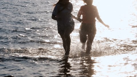 SLOW MOTION TIME WARP, SUN FLARE, CLOSE UP: Fit training partners run in refreshing sea water towards beautiful summer sunset. Glassy drops of water fly as young couple jogs through serene nature.