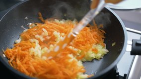 Frying chopped onions and carrots in preheated oil in pan in the kitchen at home. Slices of chopped light onion and grated carrots are mixed with silicone spatula. Cooking at home, healthy eating
