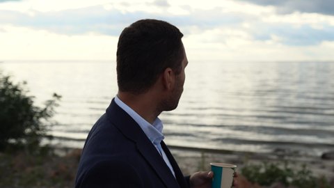 A business man stands near his car on the coast of a lake, sea or ocean, drinks coffee and eats a roll