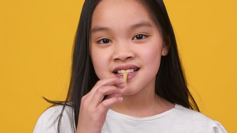 Kids eating habits. Close up portrait of adorable little asian girl eating tasty french fries, enjoying fast food, looking at camera over orange studio background, slow motion
