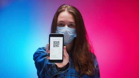 Woman wearing medical mask showing on phone vaccination passport with qr code, international coronavirus covid 19 vaccine certificate on mobile device, green zone. Red and blue neon background.
