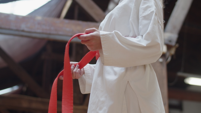 Girl tying red belt on kimono and folding arms behind back. Side view of confident female fighter preparing for karate workout in practice room. martial arts, sport concept Royalty-Free Stock Footage #1083406882