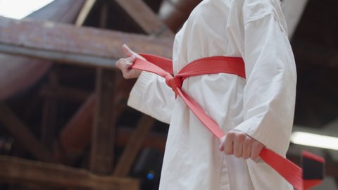 Girl tying red belt on kimono and folding arms behind back. Side view of confident female fighter preparing for karate workout in practice room. martial arts, sport concept