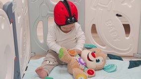 Baby boy sitting on the floor playing with soft musical educational dog toy, kid in baby safety helmet in infants playpen kids activity centre play yard. High quality 4k footage