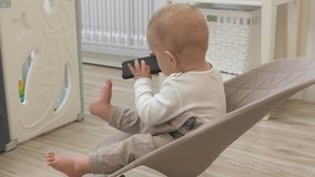 Attractive baby boy playing with mobile phone at home, cute kid using smartphone sitting in a rocking chair for babies. High quality 4k footage