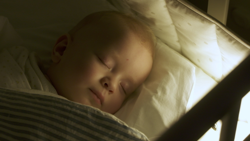 Swaddled 9 months baby boy sleeping in crib in evening in light of night lamp. Blanket is wrapped around child to calm him down and fall asleep. High quality 4k footage | Shutterstock HD Video #1083413014