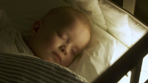 Swaddled 9 months baby boy sleeping in crib in evening in light of night lamp. Blanket is wrapped around child to calm him down and fall asleep. High quality 4k footage