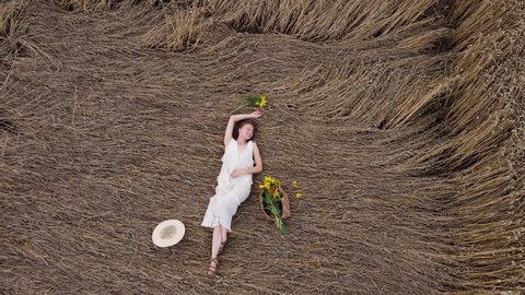 beautiful young woman in dress is lying on a wheat field. summer holiday concept. happy girl in wicker hat and bag in hand with sunflowers on nature. 4k.