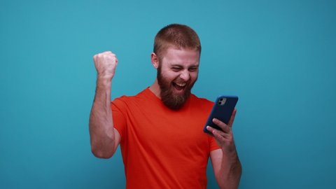 Astonished young bearded man reading shocking post on social network using cell phone, chatting looking surprised, wearing orange T-shirt. Indoor studio shot isolated on blue background.