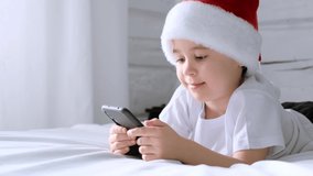 Smiling 5 year boy play smartphone video game. Said wow. Child in Santa Claus hat lie on bed. White home bedroom. Kid use mobile phone for gaming or learning. Spend time speaking with friends online.