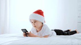 Happy 5 year boy play smartphone video game. Smiling child in Santa Claus hat lie on bed. Kid use mobile phone for gaming, learning. Online education. White home bedroom. Close-up. 4K video footage.
