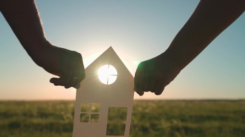 The family's hands are holding a paper house at sunset, the sun is shining through the window. The symbol of the house, happiness. The concept of building a house for the family. Dream to buy a house