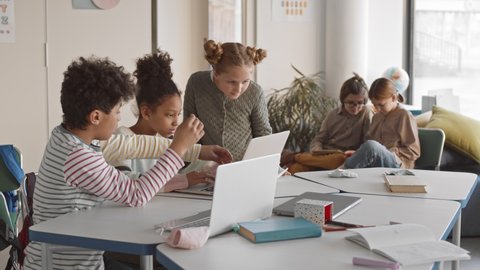 Medium slowmo of group of multiethnic schoolkids playing on laptop during break in modern informal classroom