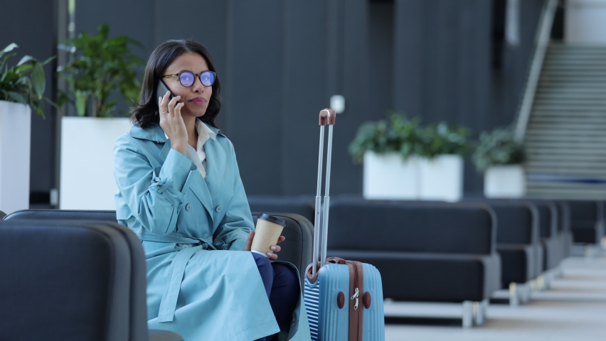 Young businesswoman having mobile call and waiting for plane while sitting in airport room spbi. Beautiful female talks on phone and looks with smile, holds coffee cup in hand and sits in lounge area