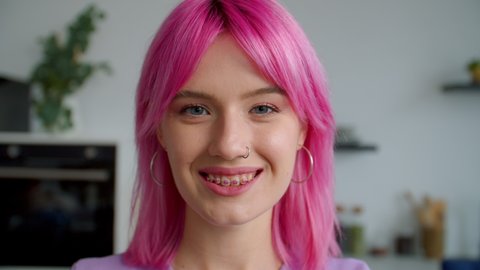 Closeup portrait of trendy beautiful pink haired young woman with dental braces flirting with camera, looking with radiant smile and enigmatic stare, expressing cheerful mood and positivity indoors.