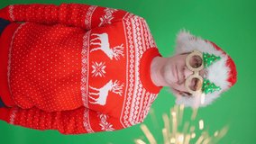 funny man in red sweater with deers wearing glasses with Christmas trees seriously looks at camera on green background with sparkles. Merry Christmas greetings vertical video. Funny New Year 2022