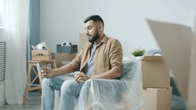 Middle Eastern guy making video call with smartphone talking showing key in new house sitting on sofa with cardboard boxes in background