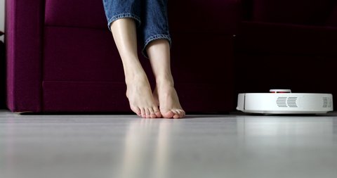 robot vacuum cleaner driving around barefoot woman at home. Female lift feet up when a smart vacuum cleaner passes to clean the floor. Concept of hygiene, household gadgets and robots at modern life