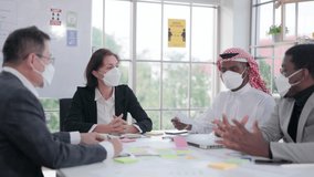 At a conference table, a multi-ethnic group of young male and female investors wearing face masks discuss business documents.