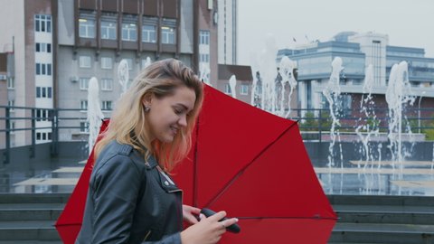 Happy girl in leather jacket walks down street past fountains, smiles coquettishly, twirls red umbrella in her hand. Attractive blonde woman enjoys every moment, flirts, is joyful of pleasant event