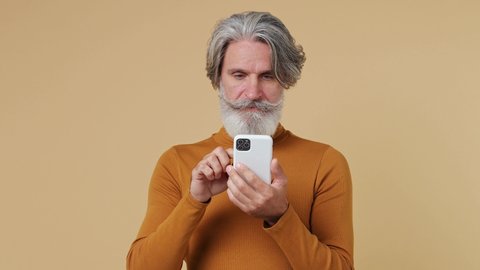 Excited cheerful amazed shocked elderly gray-haired mustache bearded man 55 years old wears brown shirt hold use point on mobile cell phone isolated on plain pastel beige background studio portrait
