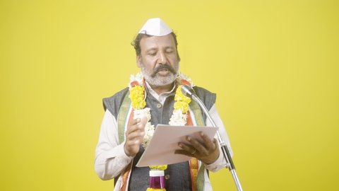 Politician seriously making oath ceremony with garland and white cap by holding papers in front of microphone on yellow background - concept of Politics, public speech or announcement.