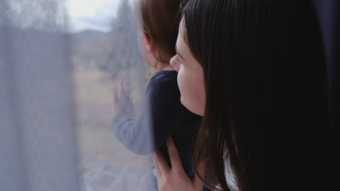 Selective focus of caring loving young mother embracing little baby girl at home. Caucasian mom and toddler daughter together standing on windowsill and looking out through window. Family love concept