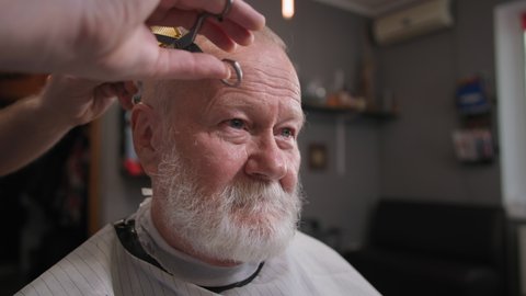 professional haircutter doing haircut with scissors and comb to stylish elderly male client in professional barbershop, close-up